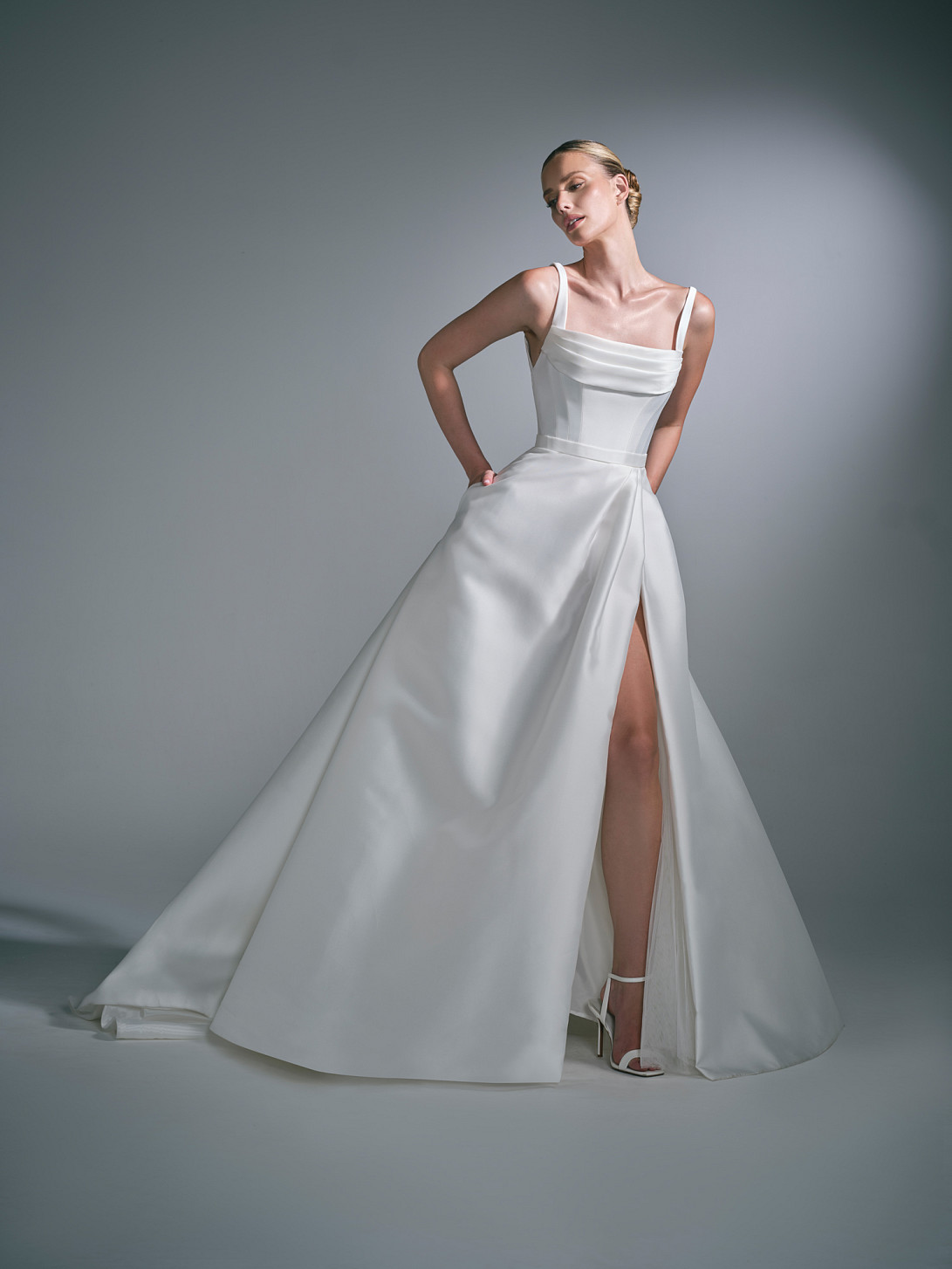 The Lysenne bridal gown has A defined waist flows into a beautifully full, softly pleated Mikado skirt, creating volume while retaining an elegant sense of movement. A high split is subtly concealed within the folds of the skirt, revealing the leg